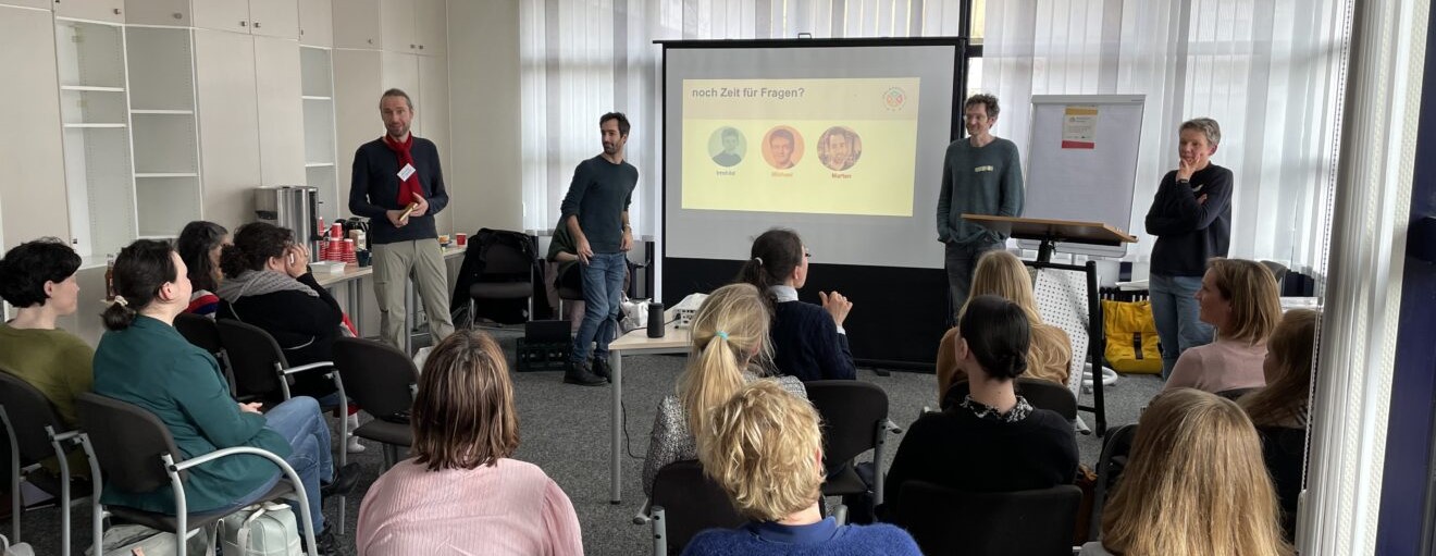 Nils, Marten und Michael von Inklupreneur stehen vor einer Leinwand vor einer Gruppe beim Startercamp. Vor ihnen sitzen einige Teilnehmende